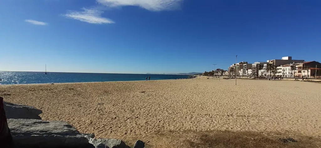 Playa de Bellamar en Premià de Mar con arena dorada, mar tranquilo y paseo marítimo al fondo en un día despejado